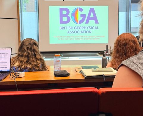 Ekranida Britaniya geofizika assotsiatsiyasi slaydi ko‘rsatilgan konferensiya zali