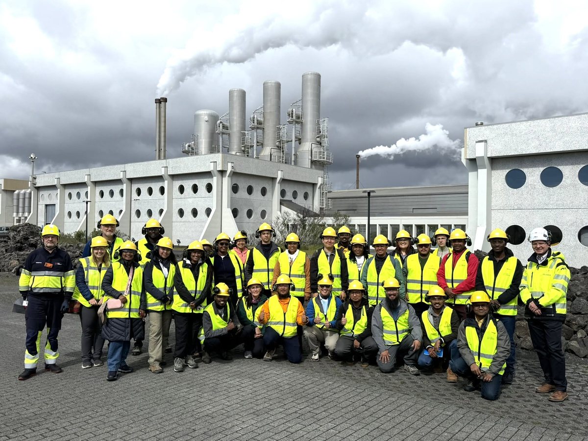 About 30 people in yellow hard hats and safety vests in front of a geothermal power plant with steaming stacks