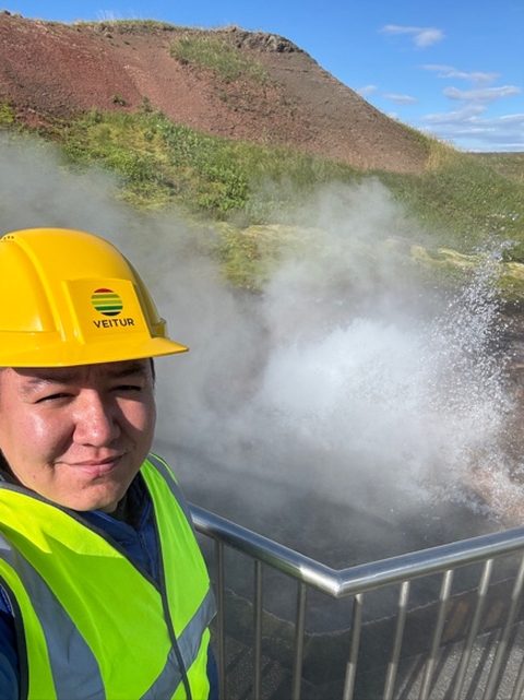 Elbek Ochilov in a hard hat and safety vest beside the steaming Deildartunguhver hot spring