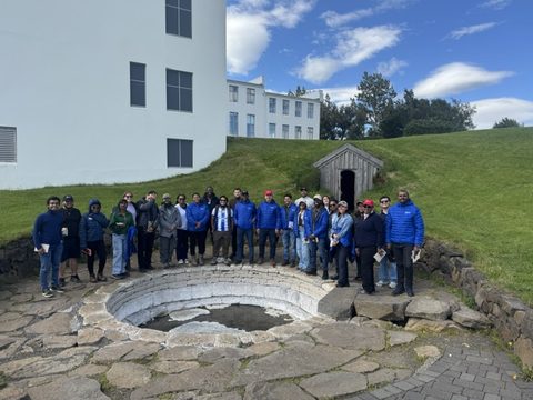 About 30 fellows standing around a round stone hot pool beside a white building